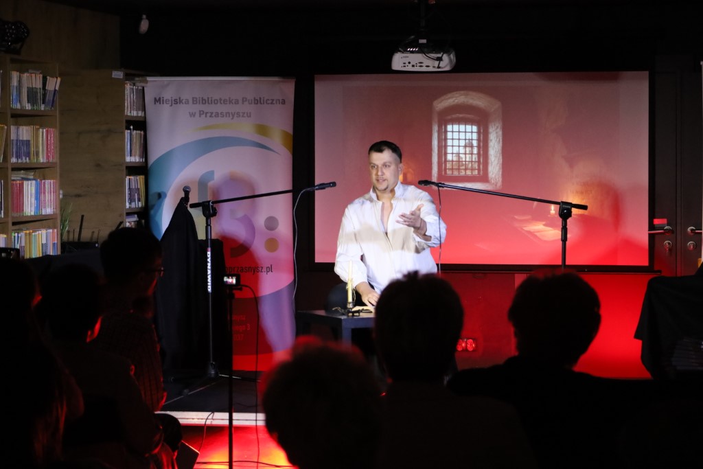 Aktor w białej koszuli występuje na scenie przed mikrofonem, gestykulując dłonią. W tle widoczny jest ekran projekcyjny z ilustracją przedstawiającą okno z kratą oraz roll-up Miejskiej Biblioteki Publicznej w Przasnyszu. Na pierwszym planie widoczne są sylwetki zgromadzonej publiczności