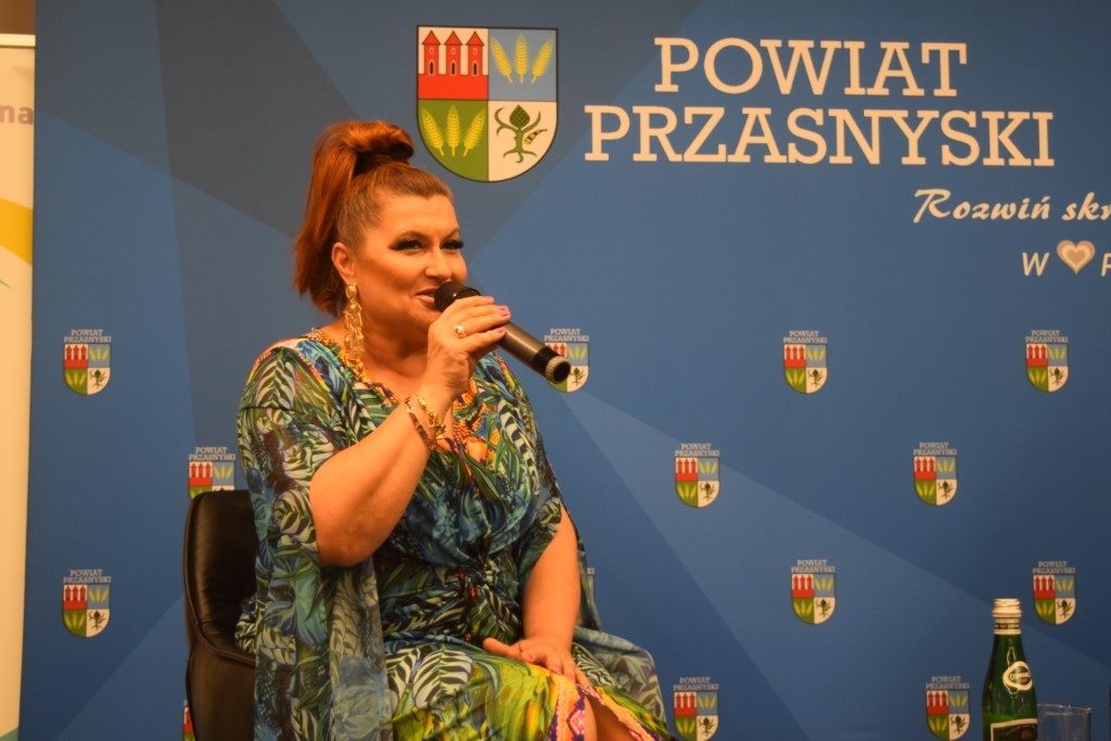 Bliskie ujęcie na uśmiechniętą Katarzynę Skrzynecką w kolorowej kreacji, która trzyma mikrofon i mówi do zgromadzonej publiczności. W tle widoczne logo Powiatu Przasnyskiego
