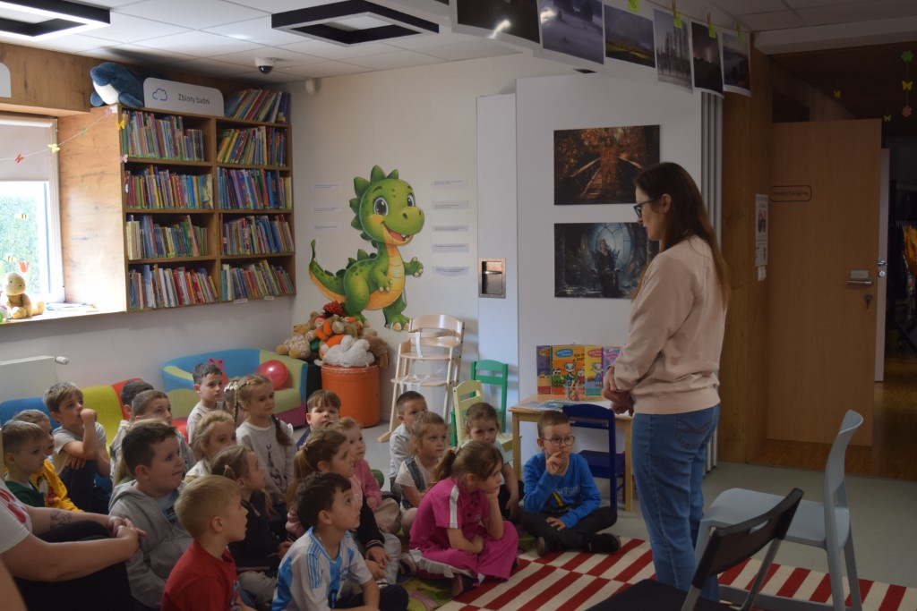 Kobieta w okularach i jasnej bluzie stoi przed grupą przedszkolaków siedzących na pasiastym dywanie w bibliotece i rozmawia z nimi. Dzieci z uwagą słuchają i patrzą w jej stronę. W tle widoczny jest kącik dziecięcy z regałami pełnymi książek, dużą naklejką przedstawiającą zielonego smoka na ścianie oraz niskimi, kolorowymi krzesełkami