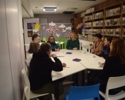 Widok na salę biblioteczną, w której osiem osób uczestniczy w spotkaniu przy wspólnym stole. Część uczestników pije napoje z białych filiżanek, na stole leżą też drobne poczęstunki w fioletowych pojemnikach. Ściany pomieszczenia zdobią półki z książkami oraz dziecięce rysunki z napisem „Wiosna”