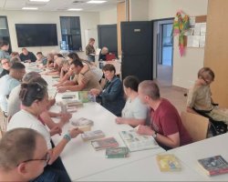 Szerokie ujęcie jaśniejszej sali bibliotecznej lub warsztatowej. Przy dwóch połączonych, białych stołach siedzi kilkunastu uczestników spotkania. Przed nimi rozłożone są liczne książki. Część osób rozmawia ze sobą, inne przeglądają strony. Po prawej stronie przy stole siedzi osoba na wózku inwalidzkim