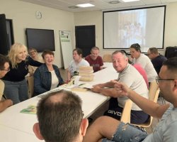 Zbliżenie na grupę rozmawiających i uśmiechniętych osób siedzących wokół białego stołu. Pośrodku stoi wysoki stos ułożonych na sobie książek. Po lewej stronie stoi blondynka w ciemnym swetrze, która uśmiecha się do siedzącej obok kobiety w jeansowej kurtce. Mężczyzna po prawej stronie wskazuje palcem w kierunku aparatu. W tle widać ekran projekcyjny