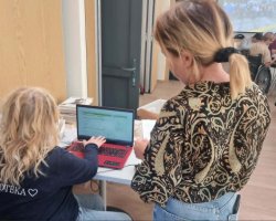 Ujęcie od tyłu. Kobieta z blond kręconymi włosami w ciemnej bluzie z napisem „BIBLIOTEKA” obsługuje czerwony laptop. Obok niej stoi druga kobieta w bluzce w zebrany wzór paisley i przygląda się ekranowi. W tle widać pozostałych uczestników spotkania siedzących w sali