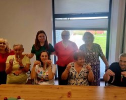 Osiem osób – pięć stojących z tyłu i trzy siedzące z przodu przy długim drewnianym stole – pozuje do zdjęcia w jasnym pomieszczeniu. Uczestnicy uśmiechają się i trzymają w dłoniach małe, ręcznie robione breloczki ze sznurka (przypominające postacie lub plecionki)