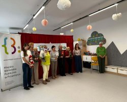 Ujęcie z boku na salę zabaw, w której grupa siedmiu kobiet stoi w rzędzie przy czerwonej kurtynie teatralnej. Część z nich trzyma w rękach lalki i pacynki. Po lewej stronie wyraźnie widoczny jest duży, biały stojak reklamowy (roll-up) z logotypem i napisem „Miejska Biblioteka Publiczna w Przasnyszu”. Pod sufitem wiszą papierowe, ozdobne kule