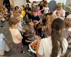 Kadrowane od góry ujęcie w sali zabaw. Na pierwszym planie widać tył głowy i ramiona kobiety trzymającej biały, wiklinowy koszyk wypełniony wielobarwnymi maskotkami z włóczki. Obok stoi mała dziewczynka w jasnej bluzce, uważnie patrząc na zawartość kosza. W tle na dywanie siedzi i bawi się grupa maluchów pod opieką dorosłych kobiet