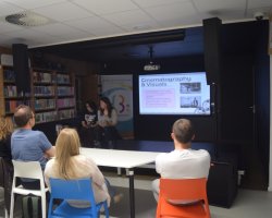 Uczestnicy Kawiarenki Językowej tyłem do obiektywu, skupieni na prezentacji multimedialnej w sali bibliotecznej. Na dużym ekranie w głębi wyświetlany jest slajd o tytule „Cinematography & Visuals” (Kinematografia i oprawa wizualna) zawierający angielski tekst oraz dwa czarno-białe kadry filmowe. Na scenie obok ekranu siedzą dwie rozmawiające kobiety prowadzące spotkanie