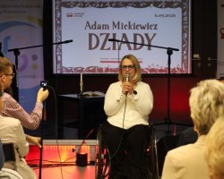 Kobieta w okularach i jasnej bluzce przemawia do mikrofonu, siedząc na wózku inwalidzkim na scenie. W tle znajduje się ekran z napisem „Narodowe Czytanie. Adam Mickiewicz DZIADY. 6.09.2026” oraz roll-up biblioteki. Z przodu widoczny jest mężczyzna nagrywający wystąpienie smartfonem oraz publiczność