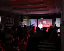 Szerokie ujęcie przyciemnionej sali widowiskowej biblioteki podczas występu. Na oświetlonej czerwonym światłem scenie występuje mężczyzna w białej koszuli. Wypełniona po brzegi sala jest widoczna z perspektywy tyłu – z wyraźnymi zarysami sylwetek siedzących widzów