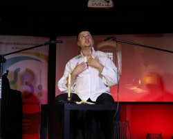 Mężczyzna w białej rozpiętej koszuli siedzi na podwyższeniu na scenie, trzyma dłoń na piersi i patrzy w górę w ekspresyjnej geście aktorskim. Obok niego na stoliku stoi zapalona świeca oraz otwarta książka. W tle znajduje się ekran z projekcją i mikrofon na statywie