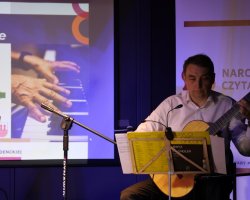 Mężczyzna w jasnej koszuli gra na gitarze klasycznej, siedząc przed pulpitem z nutami. W tle widoczny jest ekran projekcyjny ze slajdem prezentacji oraz fragment napisu „Narodowe Czytanie”
