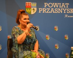 Bliskie ujęcie na uśmiechniętą Katarzynę Skrzynecką w kolorowej kreacji, która trzyma mikrofon i mówi do zgromadzonej publiczności. W tle widoczne logo Powiatu Przasnyskiego