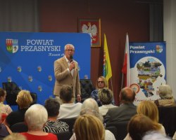 Mężczyzna w beżowej marynarce przemawia do mikrofonu przed widownią. W tle niebieska ścianka z logo Powiatu Przasnyskiego oraz polskie flagi
