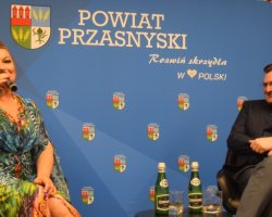 Katarzyna Skrzynecka mówi do mikrofonu, siedząc na fotelu obok prowadzącego w ciemnej marynarce, który z uśmiechem jej słucha. Na małym stoliku przed nimi stoją butelki z wodą