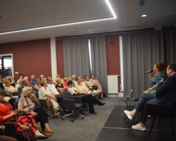 Widok z boku na pełną salę widzów siedzących na krzesłach oraz umieszczoną na podwyższeniu scenę, na której Katarzyna Skrzynecka rozmawia z prowadzącym spotkanie