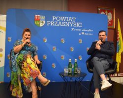 Katarzyna Skrzynecka i prowadzący spotkanie siedzą w czarnych fotelach na scenie. Oboje trzymają mikrofony i uśmiechają się, prowadząc dialog