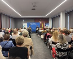 Szerokie ujęcie z perspektywy tyłu sali na licznie przybyłych uczestników spotkania, przysłuchujących się gościom na scenie pod niebieską ścianką Powiatu Przasnyskiego