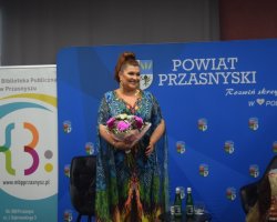 Katarzyna Skrzynecka w kolorowej, wzorzystej sukni trzyma bukiet kwiatów i uśmiecha się do publiczności na tle niebieskiej ścianki promocyjnej Powiatu Przasnyskiego i Miejskiej Biblioteki Publicznej w Przasnyszu