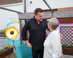 Robert Kudelski rozmawia z bliska ze starszą kobietą na scenie. Artysta nachyla się z ciepłym uśmiechem w stronę uczestniczki spotkania, trzymając dłoń na jej ramieniu. Kobieta ma krótkie, siwe włosy, nosi okulary i jasną, ażurową bluzkę. W tle stoi złota lampa sceniczna z włączonym światłem