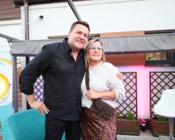 Robert Kudelski w czarnej koszuli pozuje do zdjęcia z kobietą w okularach, kremowym swetrze i spódnicy w panterkę. Artysta opiera jedną dłoń na biodrze, a drugą ręką obejmuje kobietę. Oboje uśmiechają się w stronę obiektywu. W tle widoczne są turkusowy fotel, złota lampa oraz oświetlenie sceniczne