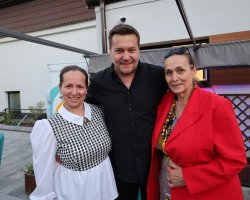 Pamiątkowa fotografia Roberta Kudelskiego stojącego pomiędzy dwiema kobietami. Po jego prawej stronie stoi kobieta w białej bluzce z wzorem w czarno-białą jodełkę, a po lewej kobieta w wyrazistej, czerwonej marynarce z okularami przeciwsłonecznymi na głowie. Wszyscy troje uśmiechają się szeroko do aparatu na tle plenerowej sceny