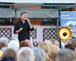 Robert Kudelski śpiewa na scenie pod zadaszeniem z materiału. Artysta uśmiecha się, trzymając mikrofon w prawej ręce i delikatnie gestykulując drugą dłonią. Za jego plecami znajduje się baner promocyjny Powiatu Przasnyskiego ze zdjęciami zabytków i krajobrazów. Pierwszy plan zajmują głowy widzów wsłuchanych w występ