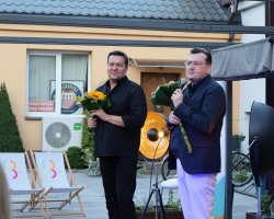 Robert Kudelski oraz prowadzący spotkanie stoją na scenie po zakończeniu występu, trzymając w rękach bukiety żółtych słoneczników. Kudelski, w czarnej koszuli, uśmiecha się łagodnie w stronę widowni. Prowadzący w ciemnogranatowej marynarce, jasnych spodniach i okularach przemawia do mikrofonu. W tle widoczny jest fragment budynku z godłem i napisem „Ochotnicza Straż Pożarna” oraz leżaki biblioteczne