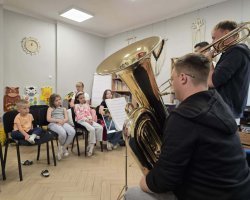 Dzieci siedzą na krzesłach i z zaciekawieniem słuchają dwóch muzyków grających na tubie i puzonie. Widok z boku na grających instrumentalistów i uśmiechniętą widownię