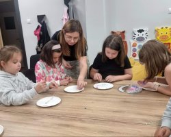 Grupa dzieci siedzi przy drewnianym stole i w skupieniu nawleka kolorowe koraliki z talerzyków na sznurki. Kobieta w okularach pochyla się nad nimi, pomagając jednej z dziewczynek