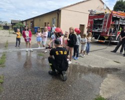 Pokaz strażacki na świeżym powietrzu. Przyklęknięty strażak pomaga dziewczynce w czerwonym hełmie trzymać prądownicę i kierować strumień wody. Wokół stoją i przyglądają się pozostałe dzieci