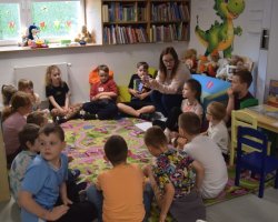 Dzieci siedzą w kręgu na wzorzystym, kolorowym dywanie w kąciku dziecięcym w bibliotece. Przed nimi kuca kobieta w okularach, która trzyma w dłoniach małą kartkę i pokazuje ją grupie. W tle widać niskie regały z książkami, pluszowe zabawki na parapecie okna oraz dużą naklejkę z zielonym smokiem na ścianie