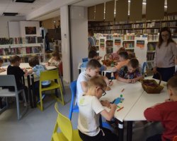 Widok na salę biblioteczną, w której dzieci pracują przy trzech różnych, wielokątnych stołach. Każda grupa wykonuje zadania plastyczne za pomocą kolorowych pisaków i kredek. Między stołami stoi kobieta w okularach, koordynująca przebieg zajęć. W tle widać długie rzędy regałów bibliotecznych oraz wiszące pod sufitem tabliczki informacyjne