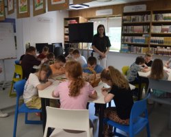 Grupa dzieci siedzi przy białych, modułowych stołach w jasnej sali bibliotecznej i wykonuje prace plastyczne. Dzieci pochylają się nad stołami, rysując pisakami i kredkami. Pośrodku jednego ze stołów stoi wiklinowy koszyk. W tle stoją dwie dorosłe kobiety, które obserwują pracę uczestników. Z prawej strony widoczne są wysokie regały ścienne z lekturami i książkami dla młodzieży